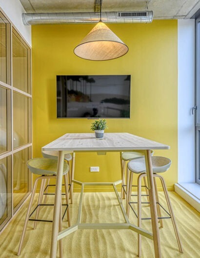 un espace de travail, une bulle avec une table haute, des sièges hauts, un écran de tv sur le mur jaune