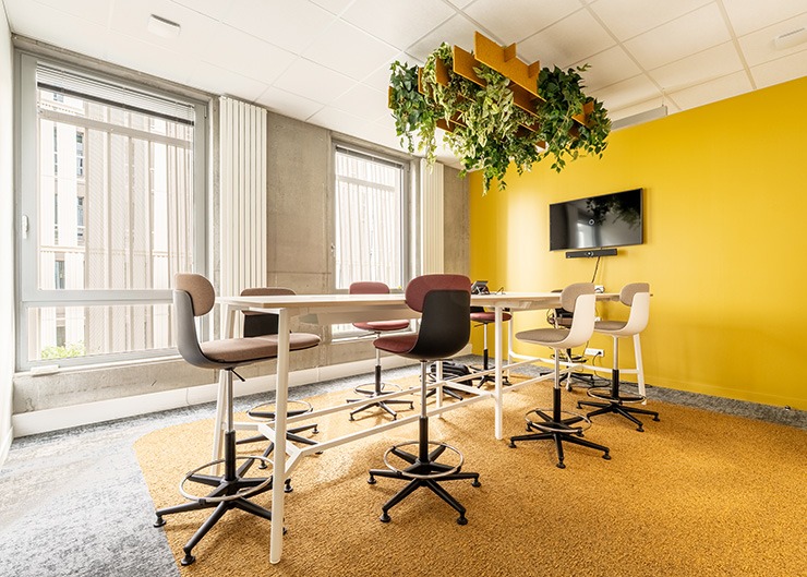 un espace collaboratif avec un mur jaune au fond, une table haute et des sièges hauts, une plante acoustique au plafond